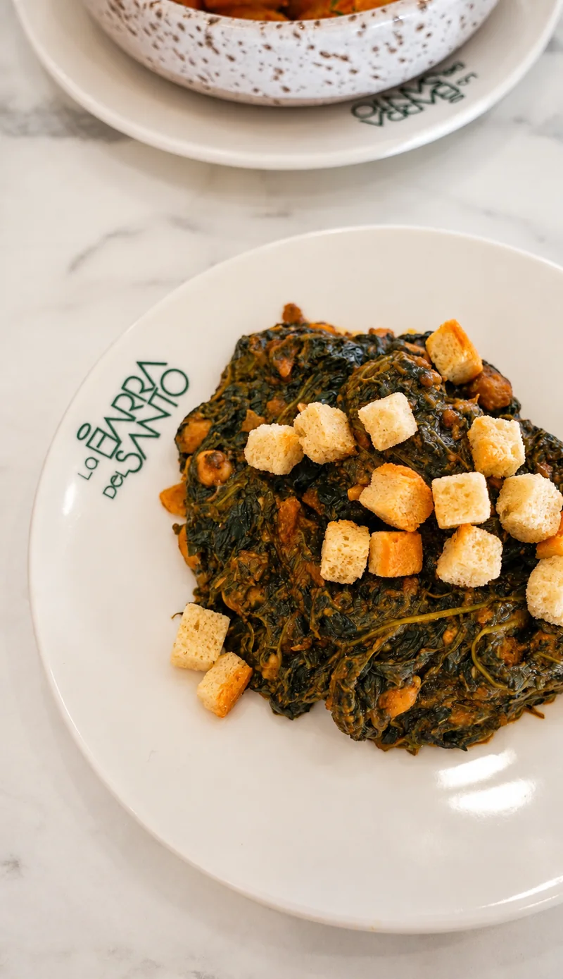 Plato con hojas verdes cocinadas y dados dorados, sobre una mesa clara.
