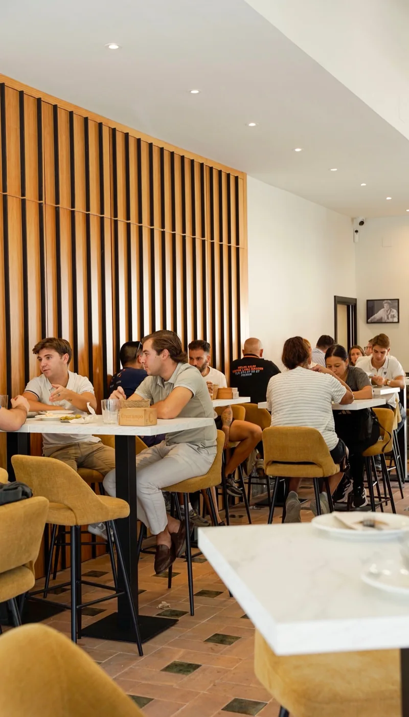 Comedor con mesas ocupadas, sillas mostaza y una pared de listones de madera.