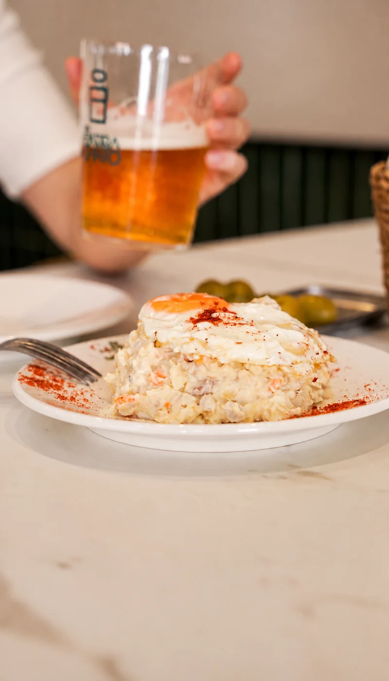 Tapa presentada en un plato blanco, con una persona sosteniendo un vaso de cerveza al fondo.